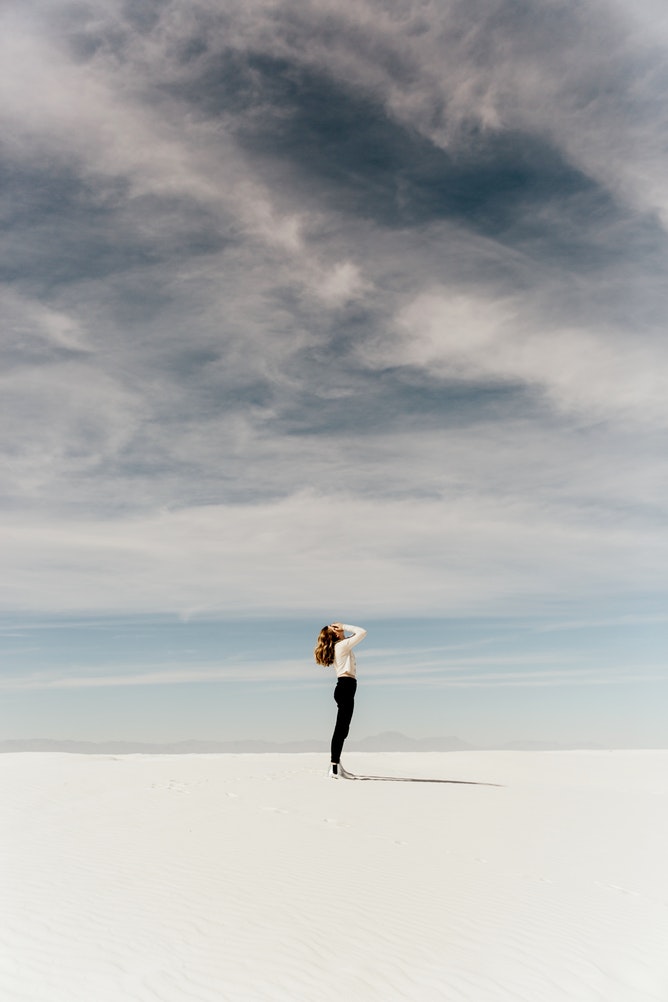 A woman in black jeans and white top holds her head in her hands while standing on salt flats
