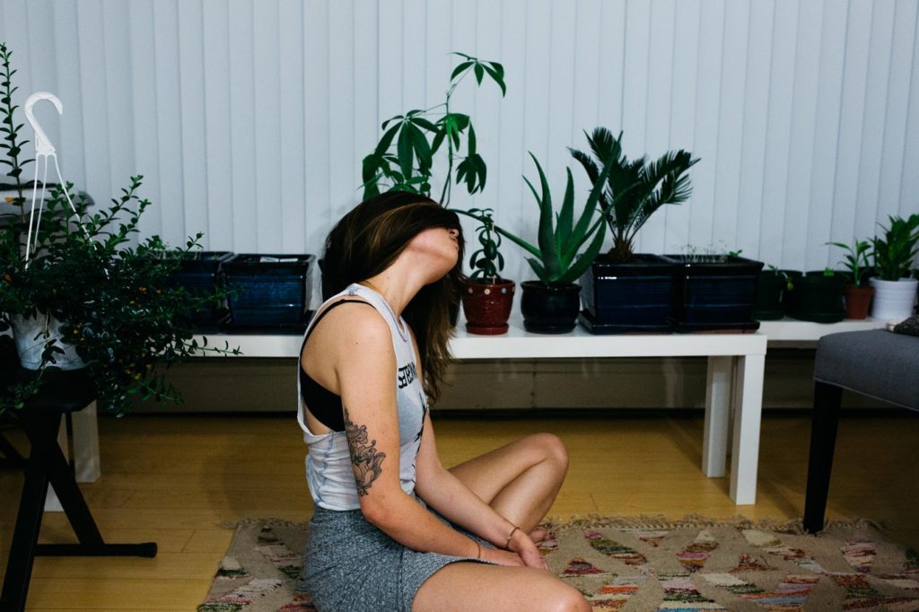 A woman with long dark hair is sitting on a rug stretching her neck