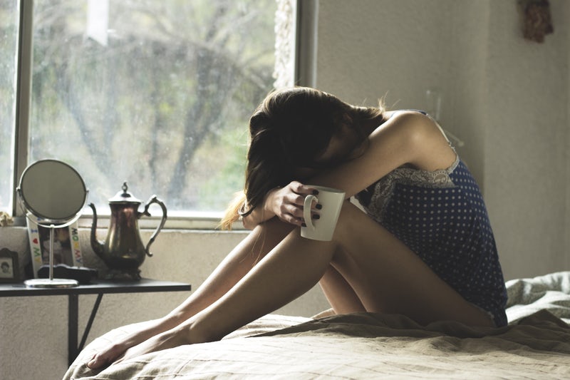 A woman, holding a cup of coffee, hangs her head in her hands