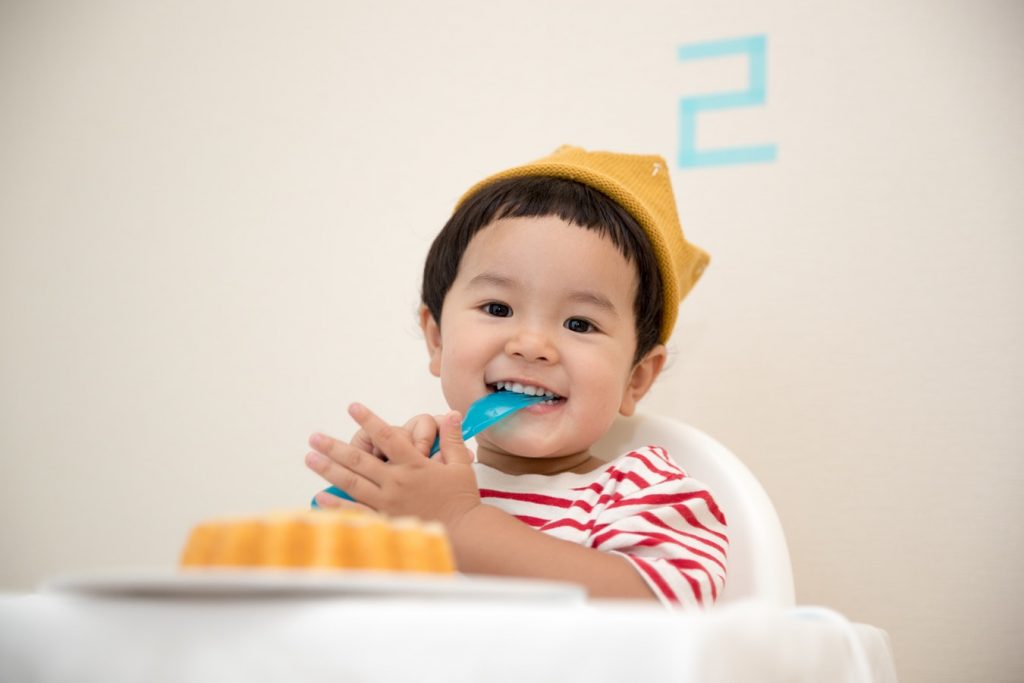 An infant with a spoon in his mouth sits in his high chair awaiting his meal